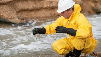 Field technician in yellow protective suit and white hard hat sampling water from a fast-flowing rocky stream